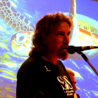José Henrique Becker falando em um microfone com uma tartaruga marinha no fundo