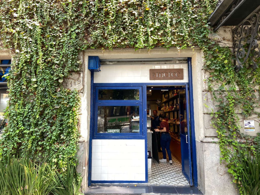 Entrada do restaurante à Mercê. Faixada tem uma porta com detalhes azuis e em volta uma planta