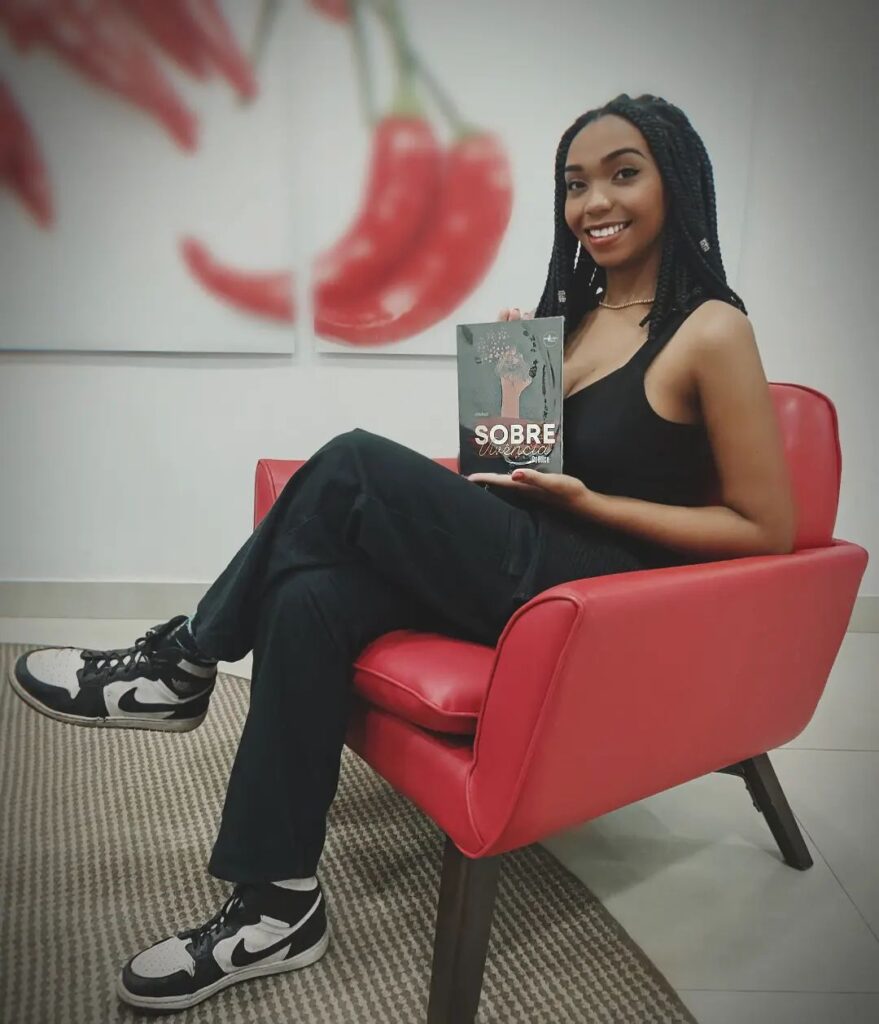 Sentada em um cadeira vermelha, a escritora Delord, que é uma mulher negra de tranças, segura o livro Sobre-Vivência Poética