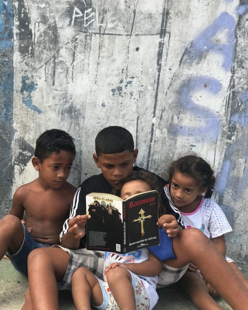 Quatro crianças sentadas no chão leeem o livro de rap Sobrevivendo no Inferno dos Racionais Mc's