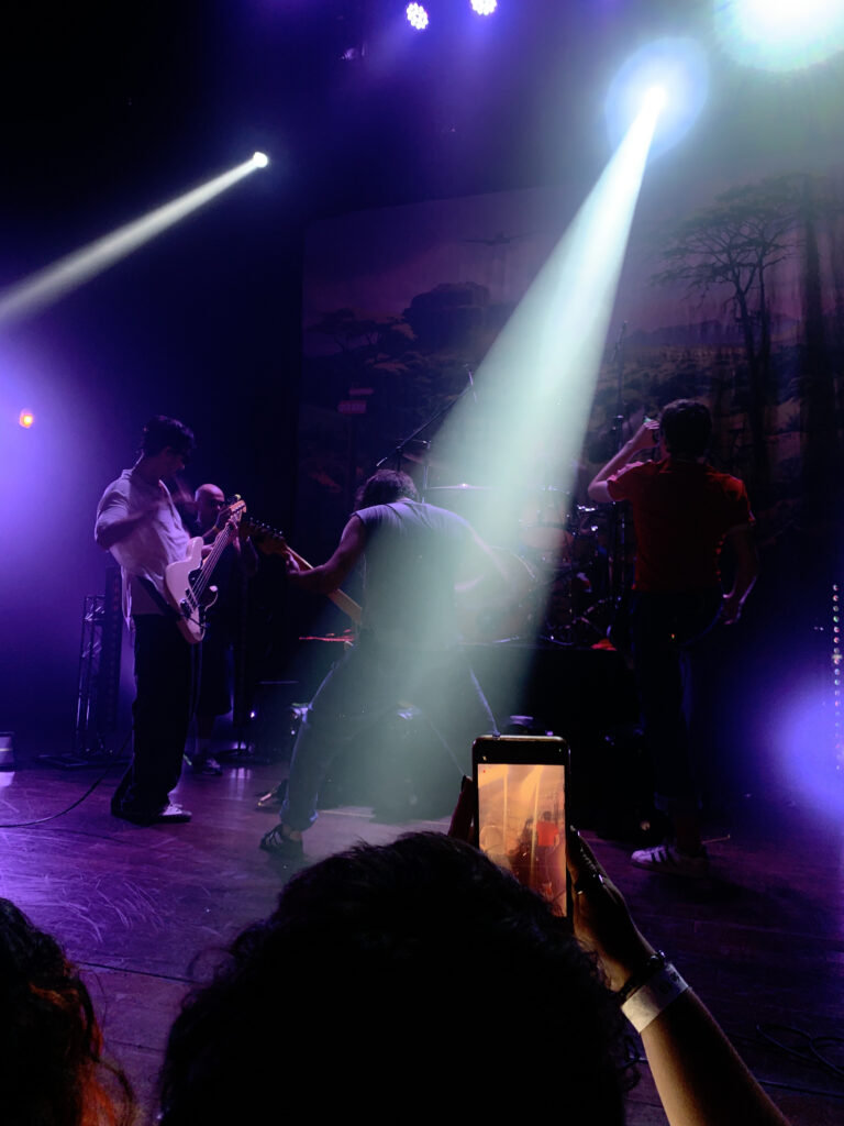 Em um palco, a Banda O Grilo performa expressivamente