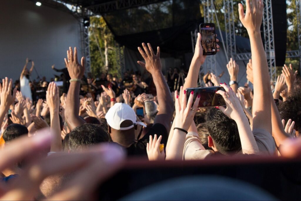 Na imagem pessoas presentes no show aparecem com os braços para cima, muitas delas segurando celulares