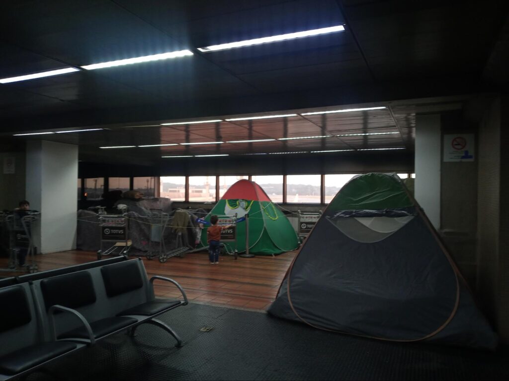 Barraca dos refugiados nas cores verde e laranja no meio do saguão do Aeroporto de Guarulhos, com crianças brincando com carrinhos de bagagem