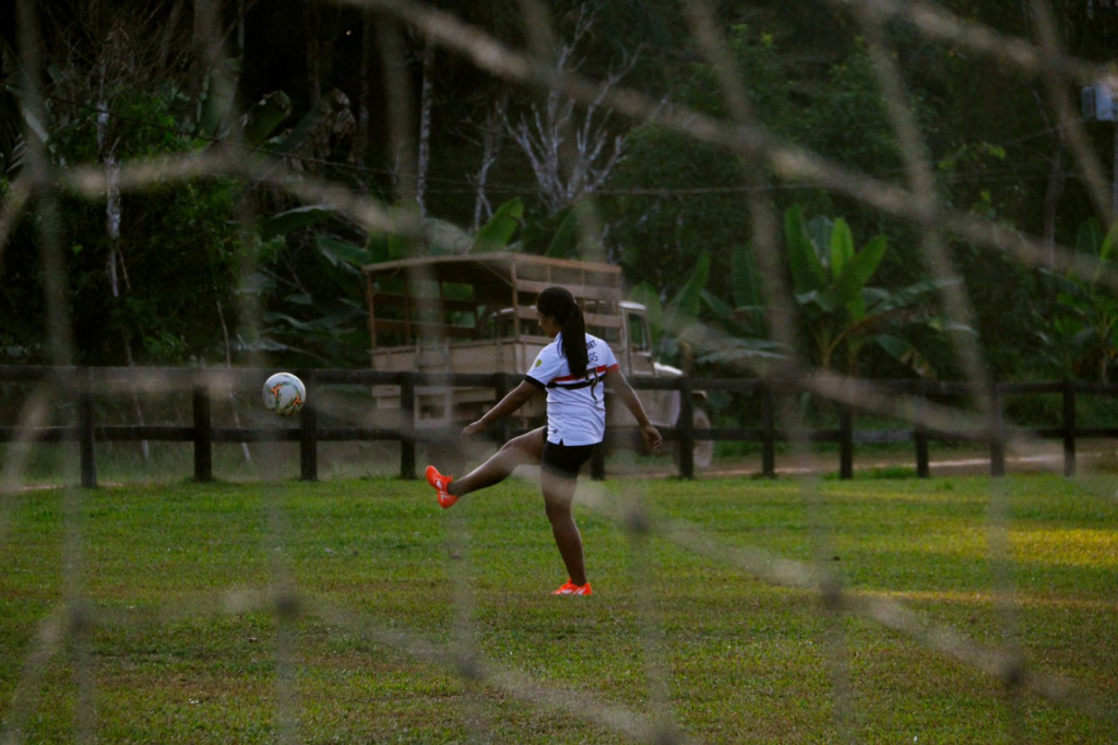 Mulher com camiseta de um time de futebol chutando uma bola para o alto. A foto foi tirada por trás de uma rede de gol, de forma que ela aparece desfocada em primeiro plano na imagem.