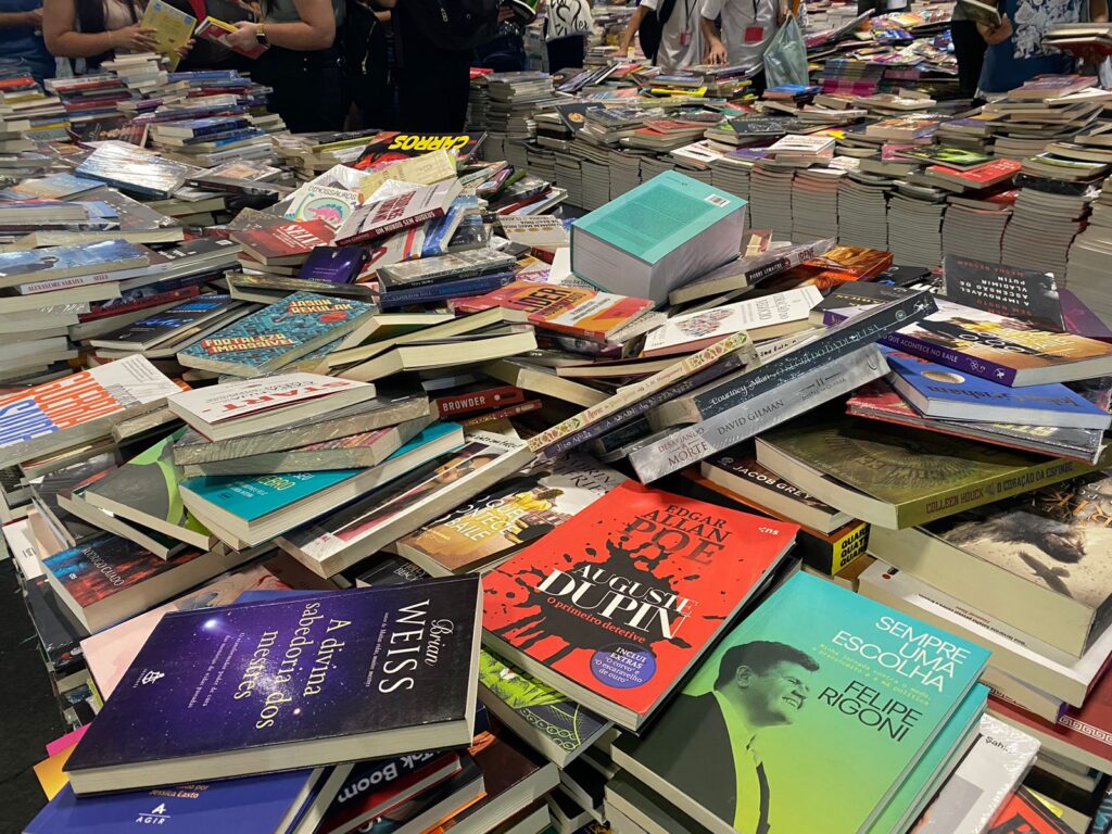 A imagem mostra um dos estandes da Bienal do Livro com vários livros empilhados desorganizadamente