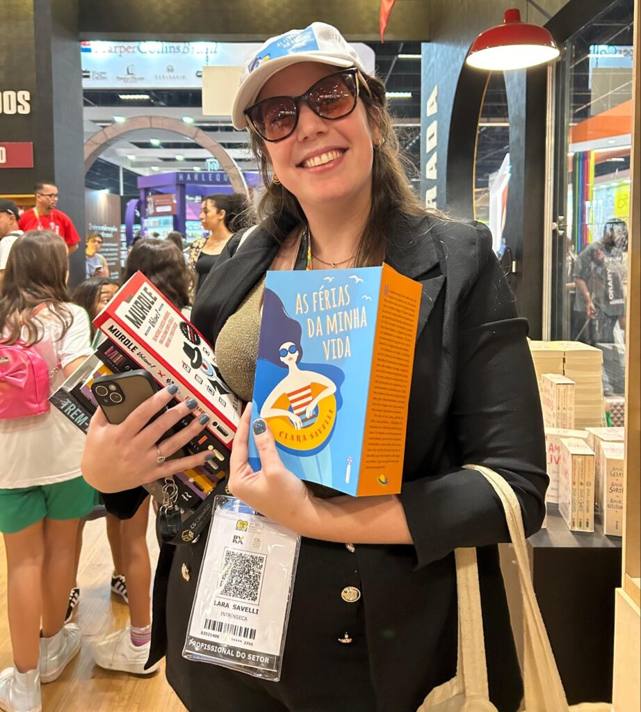 Lara Savelli com muitos livros nas mãos, destacando "As Férias da Minha Vida" na Bienal do Livro