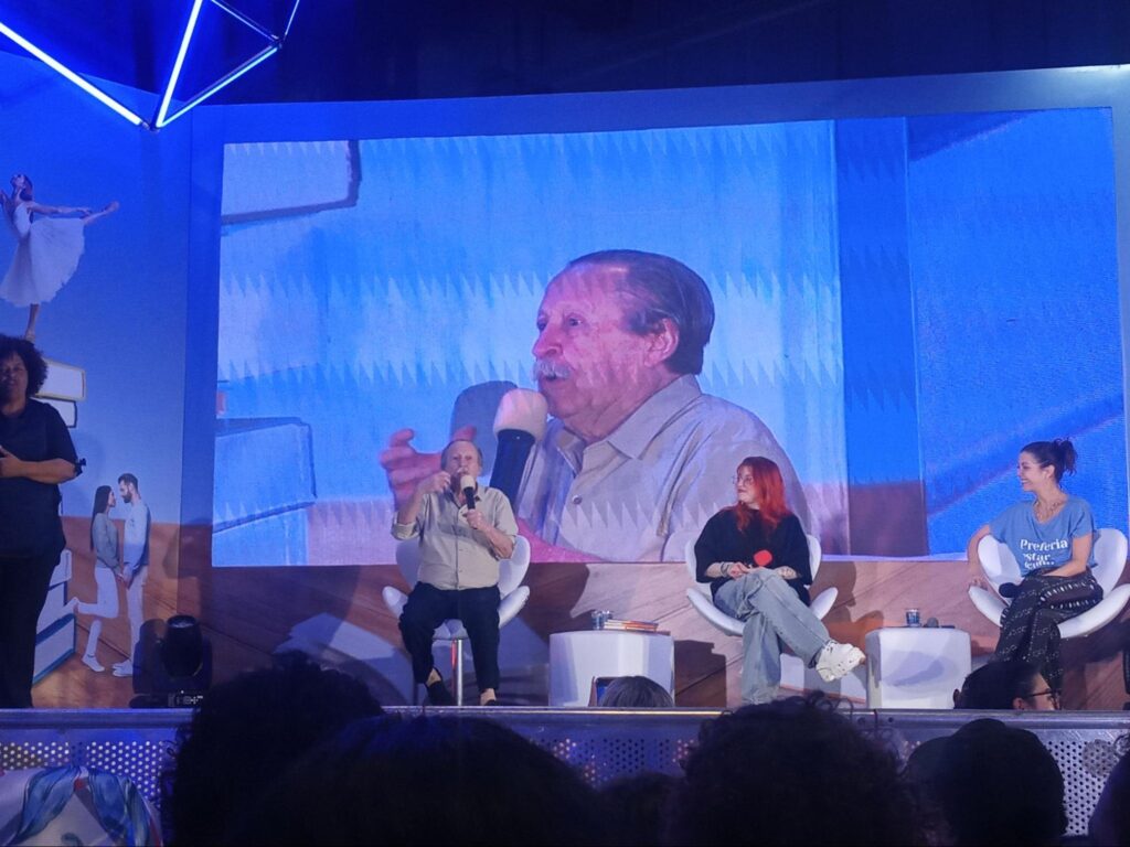 Pedro Bandeira gesticula enquanto discursa e aparece em destaque no telão do evento