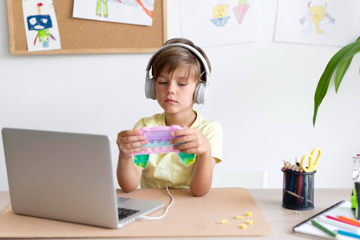 Uma criança usando fones de ouvido está sentada em uma mesa com um laptop, focada em um tablet colorido. Desenhos estão fixados na parede ao fundo.