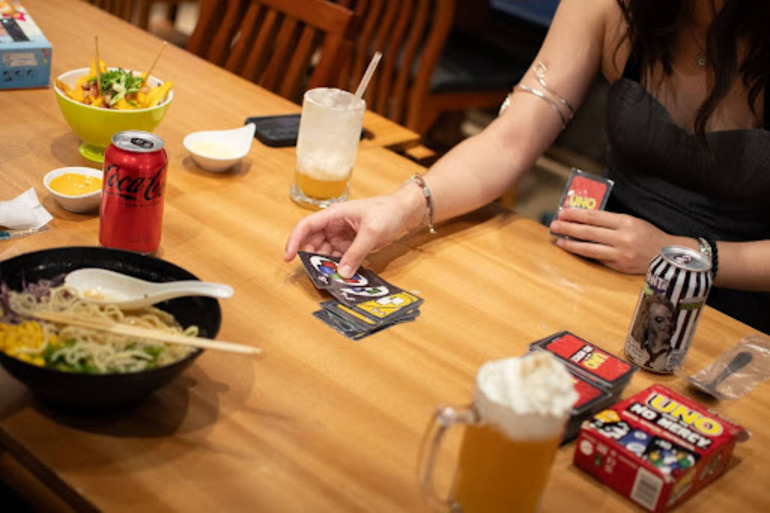 Mulher jogando um jogo de cartas enquanto come um lámen.