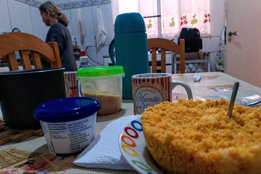 Na foto, aparece uma mesa com vários objetos em cima: um cuscuz, duas canecas, dois potes fechados, uma leiteira e uma garrafa de café. Ao fundo, há uma idosa na pia. 