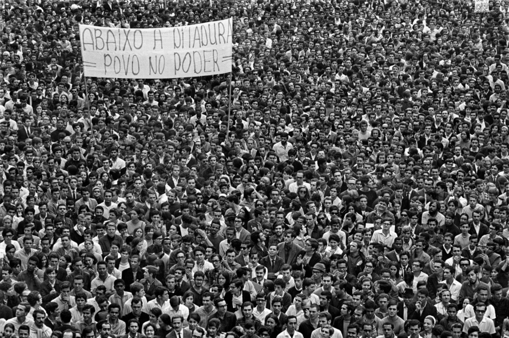 Fotografia em preto e branco de uma manifestação contrária à ditadura militar. Há uma multidão e uma faixa no canto superior esquerdo com os escritos: "Abaixo à Ditadura/Povo no poder". 