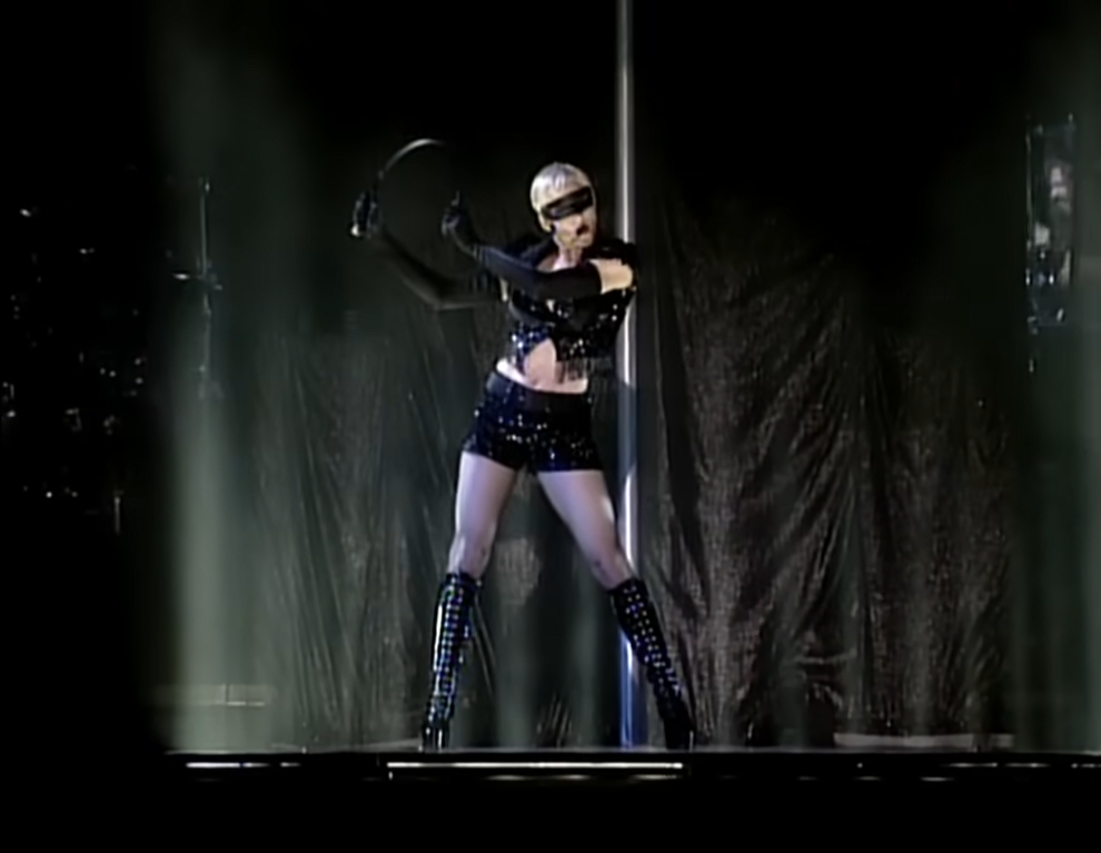 Madonna em um show. Ela usa um shorts preto colado, uma bota alta preta, um cropped preto. Todos colado em sua pele. Ela também usa uma máscara da mesma cor. 