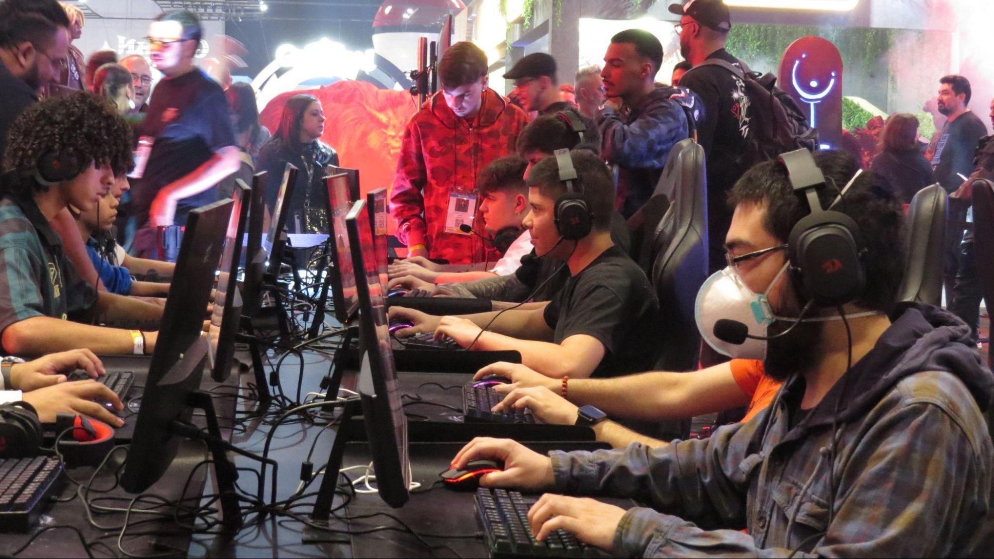 Foto lateral de uma fileira de pessoas jogando. Vamos o lado dos computadores e os braços dos jogadores, mas apenas o rosto do primeiro, que está de máscara facial e fones de ouvido