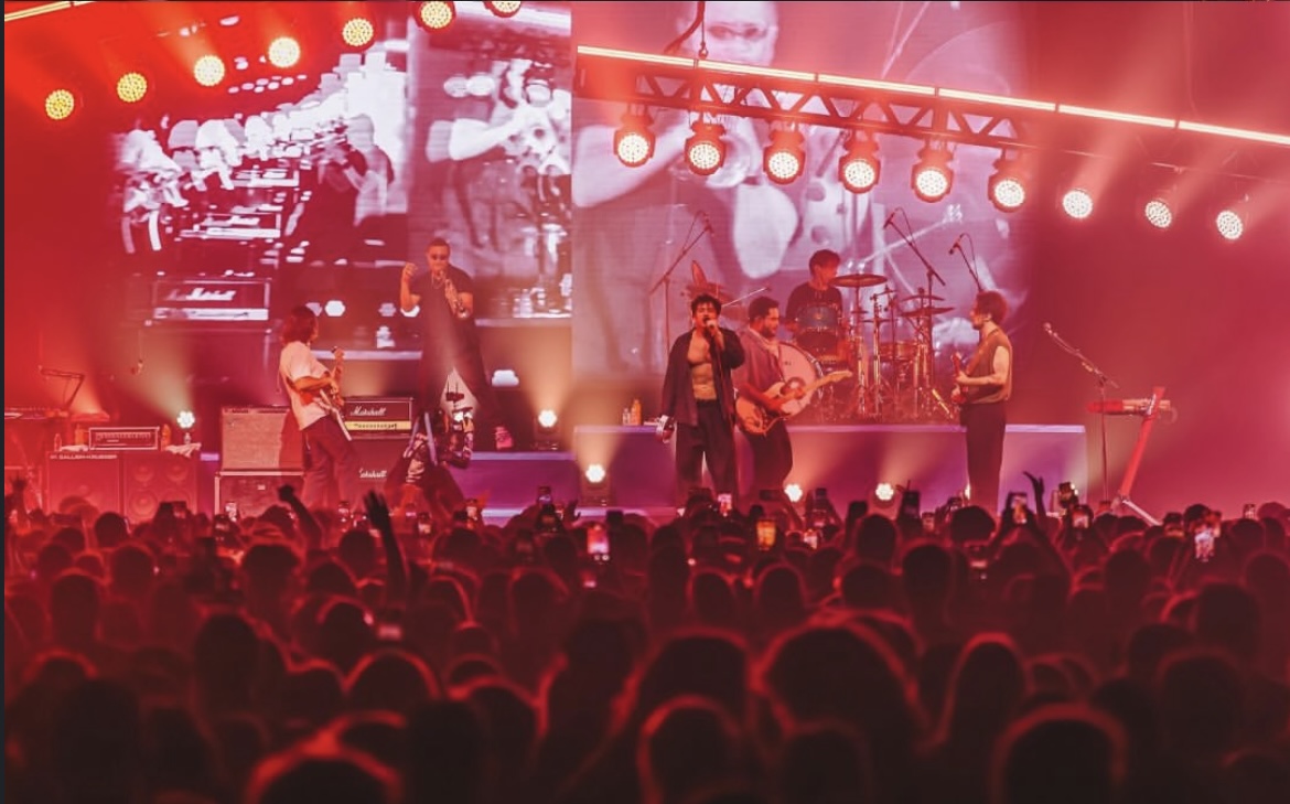 Foto do show da banda Lagum. A imagem é vermelha por conta das luzes, mostra o palco com os artistas e as cabeças da plateia