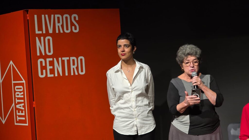 Curadora da Feira. Ao fundo, um painel vermellho escrito "Livros no centro". 