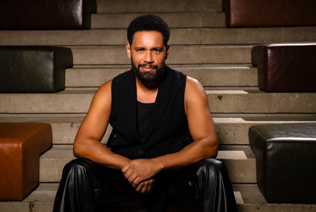 A figura apresenta César Mello, um homem negro, de barba e cabelo curto, ele está sentado e utiliza roupas pretas