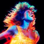 Tina Turner com cabelos ao vento e com um grande sorringo. Luzes coloridas a iluminam