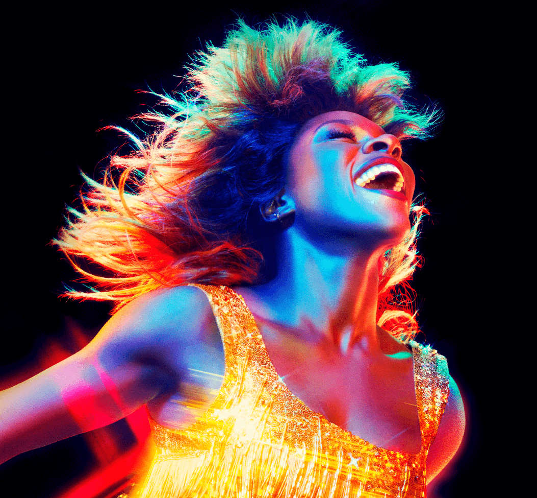Tina Turner com cabelos ao vento e com um grande sorringo. Luzes coloridas a iluminam