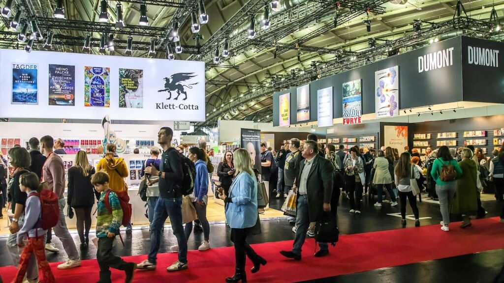 a imagem mostra a Feira do Livro de Frankfurt, evento cheio de pessoas com roupas diversas e um tapete vermelho para indicar o local onde devem passar