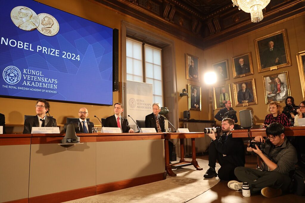 Grupo de pesquisadores reunidos em mesa recebendo prêmio Nobel com fotógrafos ao seu redor.