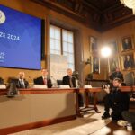 Grupo de pesquisadores reunidos em mesa recebendo prêmio Nobel com fotógrafos ao seu redor.