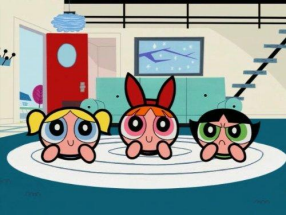 A imagem mosrra as Meninas Superpoderosas deitadas, como se estivessem assistindo televião, em um tapete branco localizado em uma sala de estar. 