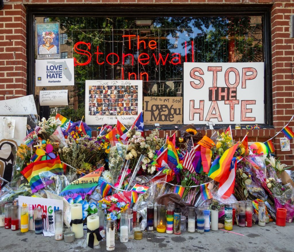 Na imagem vemos homenagens com diversas bandeiras que representam a comunidade LGBTQIAPN+ e placas com mensagens contra o ódio a pessoas LGBTQIAPN+em frente ao Stonewall Inn