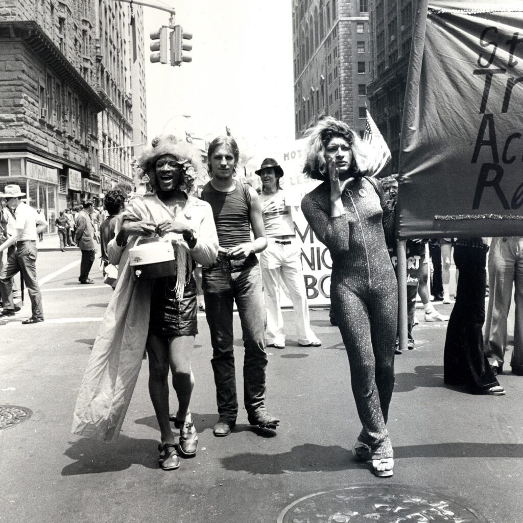 Rivera e Thompson em uma manifestação pacífica em nova York. Ambas aparecem em pé e com roupas belas, Thompson com uma mini saia, camisa e um exuberante chapéu e Rivera com macacão. Thompson possui um grande sorriso na face enquanto Rivera se mostra séria e com a mão na face enquanto segura um lado de um grande cartaz