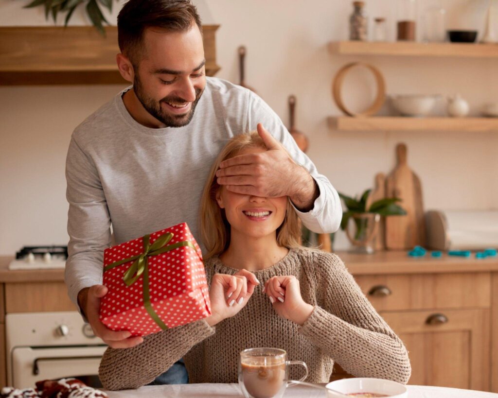 Com uma atmposfera romântica, uma mulhjer tem o rosto coberto por seu perceiro que, por sua vez, segura um presente. É perceptível que o presente se trata de uma surpresa carinhosa para a mulher