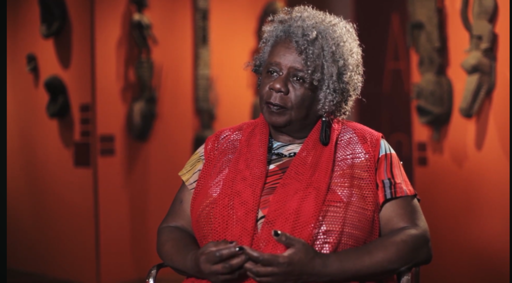 Conceição Evaristo, uma mulher negra com cabelos grizalhos e caheados vestida com uma camiseta com cores quentes e um chale vermelho, é apresentada sentada gestivulando para uma entrevista.
