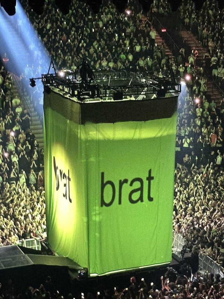 a imagem mostra um show onde grandes tendas com a capa do ábum — fundo verde-limão com tipografia sem serifa — é extendida