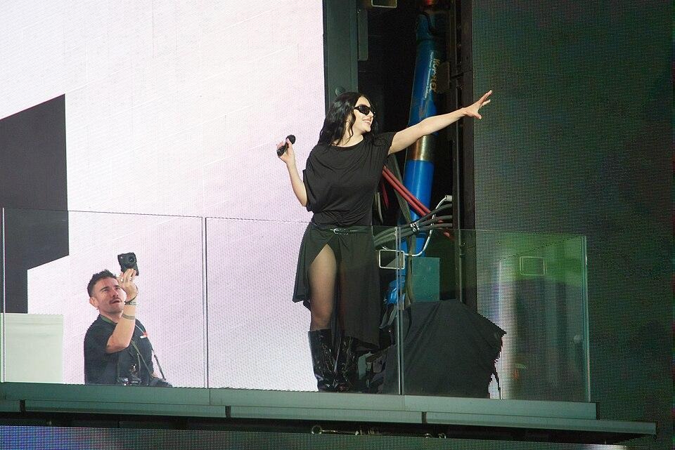 a cantora Charli XCX, uma mulher branca com cabelo preto, está em cima de um palco e veste uma roupa toda preta que consiste em um óculos de sol, botas, e um vestido com uma modelagem similar a uma camiseta, um cinto é responsável por marcar a cintura da artista