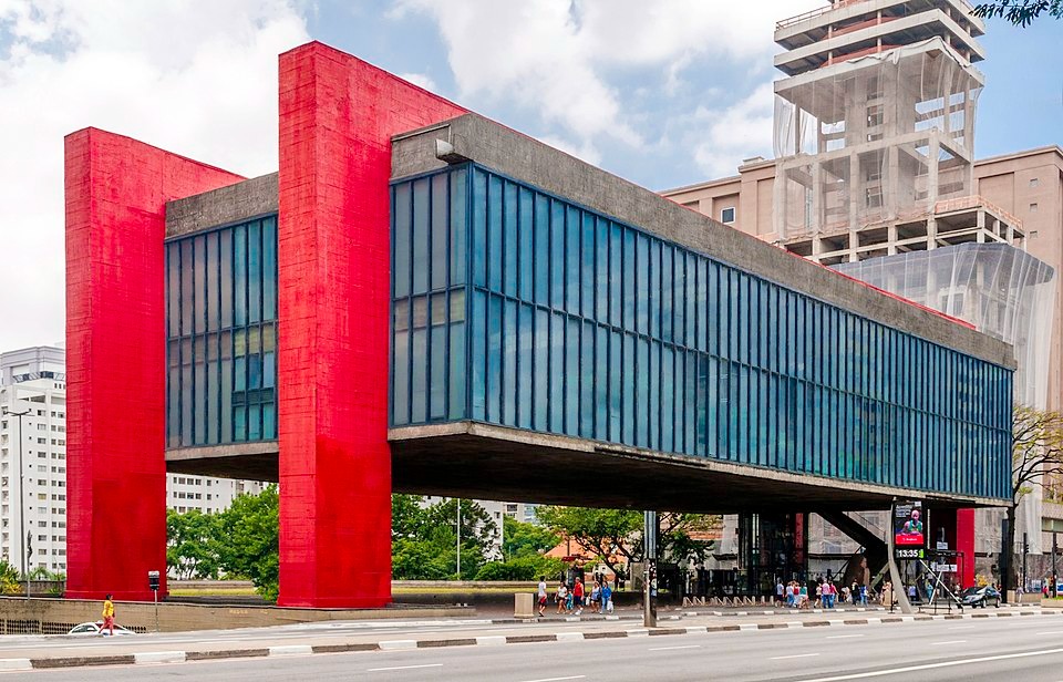 a foto mosstra o masp. um museu em formato quadrado com um grtande vão livre e vigas marcates pela sua coloração vermelha. o museu aparenta estar flutuando. a foto também mostra turistas e um baixo movimento de pedestres e veículos na avenida paulista. a iluminação é alta já que está de dia.