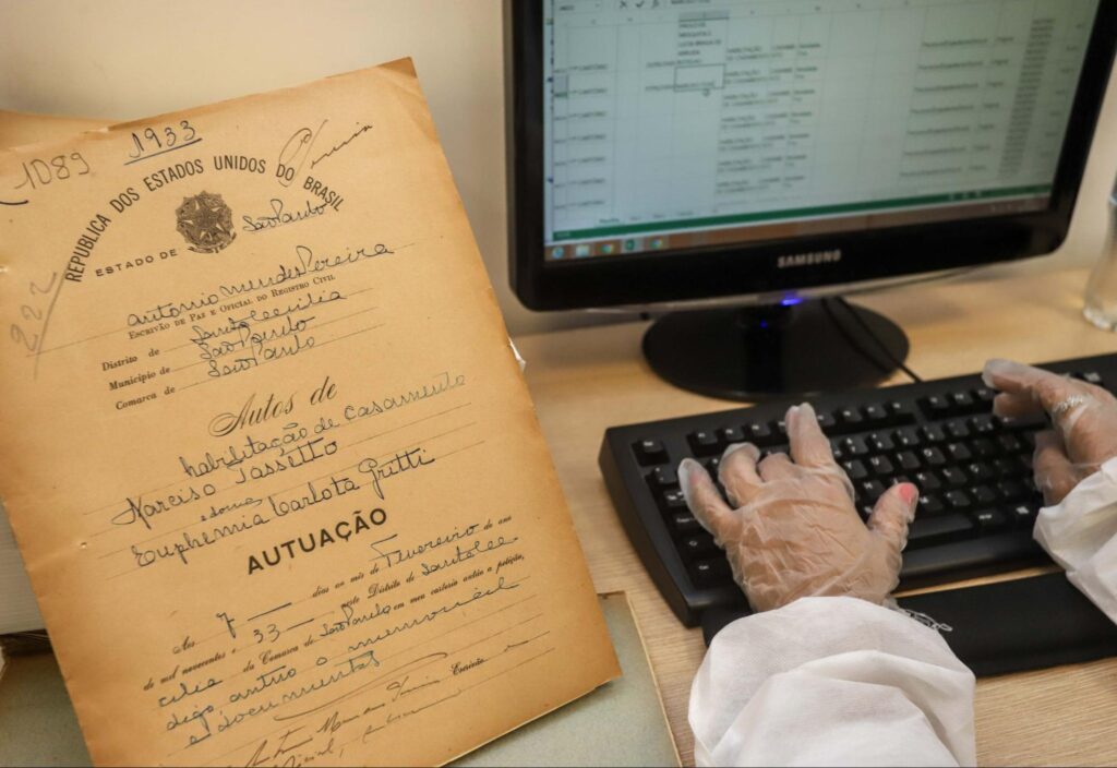 Mãos digitando em um computador informações de um documento antigo.