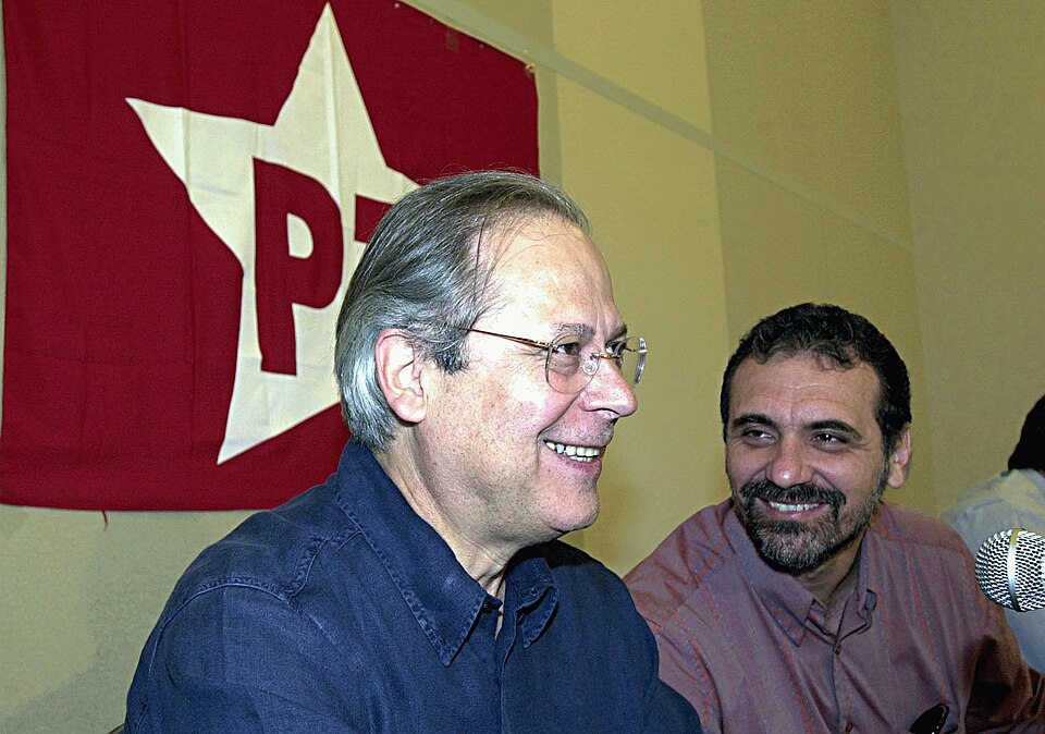 Dois homens brancos sorrindo na frente de uma bandeira vermelha