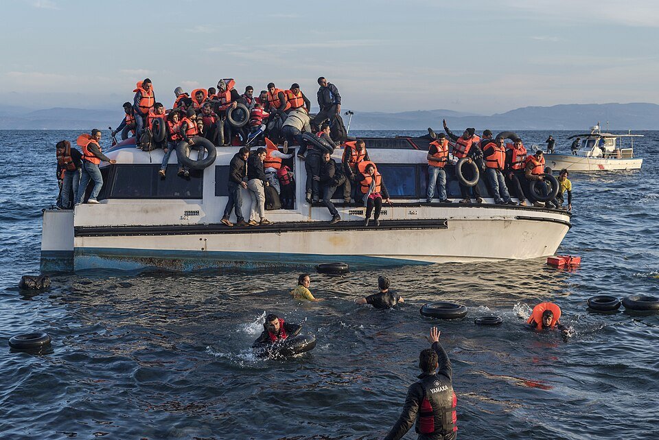 Fotografia em plano geral e iluminação natural mostrando uma embarcação lotada de refugiados no mar agitado. O barco branco, de pequeno a médio porte, carrega dezenas de pessoas amontoadas tanto no convés quanto no teto, a maioria vestindo coletes salva-vidas laranjas. Algumas pessoas pulam ou caem na água e nadam em direção à margem com a ajuda de boias e pneuzinhos pretos, enquanto em primeiro plano, de costas, uma pessoa em pé na água acena orientando os sobreviventes. Ao fundo, sob um céu claro e suave de fim de tarde, veem-se montanhas distantes no horizonte e uma pequena embarcação de resgate branca à direita.