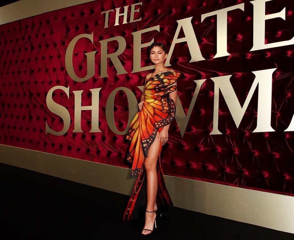 Fotografia em plano inteiro e iluminação de evento da atriz Zendaya posando no tapete vermelho. Ela veste um vestido longo e assimétrico com estampa de borboleta monarca, em tons vibrantes de laranja, preto e amarelo, com um ombro só e uma fenda profunda que exibe sua perna esquerda. A atriz está posicionada em pé no centro da imagem, usando salto alto preto fino e com os cabelos presos de forma alinhada para trás. Ao fundo, há um painel vermelho aveludado com letras grandes em dourado e branco onde se lê parcialmente a inscrição do título do filme "THE GREATEST SHOWMAN".