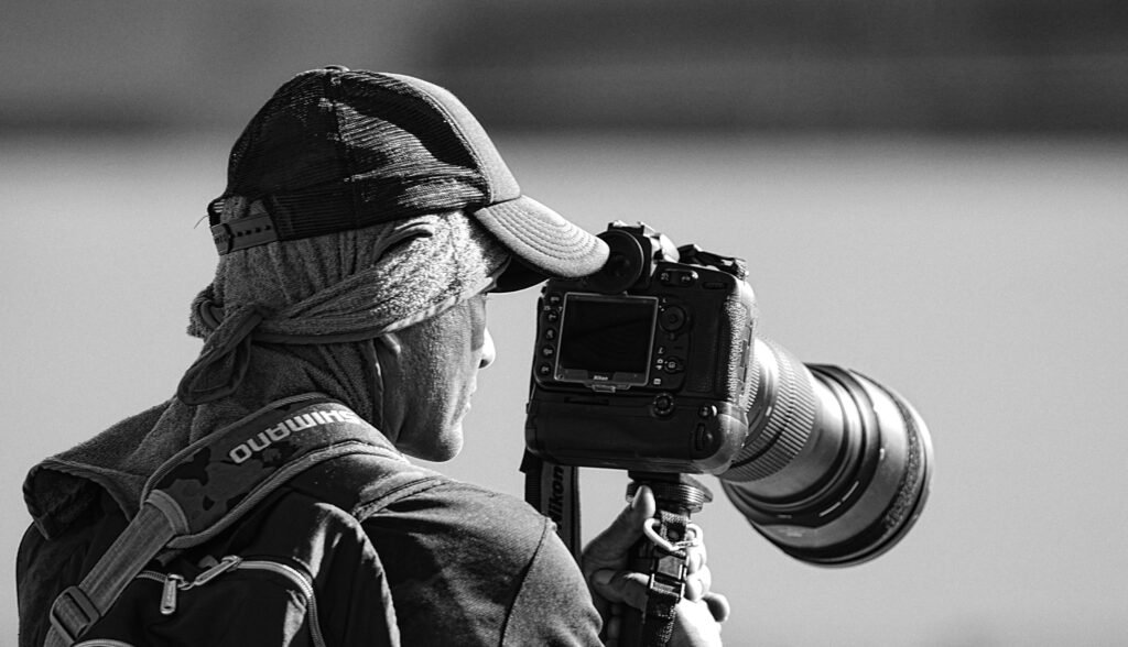 O fotógrafo é visto por trás e de lado, olhando para o visor de uma câmera profissional montada em um tripé. Ele usa um boné de beisebol com uma seção traseira de malha sobre um lenço na cabeça que está amarrado no pescoço e uma toalha enrolada no pescoço. Ele também usa uma mochila, com "SHIMANO" visível na alça direita. A câmera profissional com uma lente telefoto é montada em um tripé, que ele segura com a mão esquerda enquanto olha para o visor.