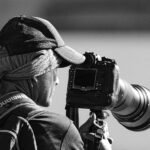 O fotógrafo é visto por trás e de lado, olhando para o visor de uma câmera profissional montada em um tripé. Ele usa um boné de beisebol com uma seção traseira de malha sobre um lenço na cabeça que está amarrado no pescoço e uma toalha enrolada no pescoço. Ele também usa uma mochila, com "SHIMANO" visível na alça direita. A câmera profissional com uma lente telefoto é montada em um tripé, que ele segura com a mão esquerda enquanto olha para o visor.