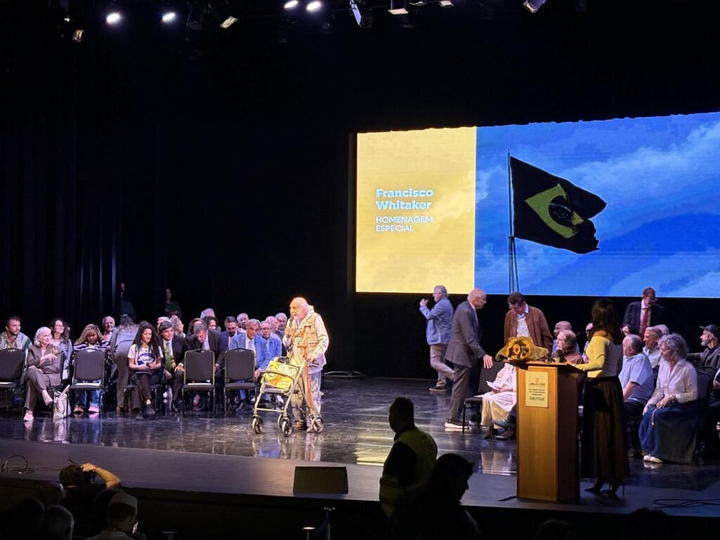 Palco preto com um telão com a bandeira do Brasil ao fundo. Na frente, um senhor branco de roupas claras