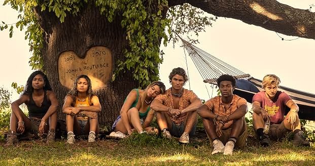 Fotografia em plano médio e em cores, com tom amarelado e quente, de seis jovens sentados em linha no chão sobre a grama, ao pé do tronco grosso de uma árvore frondosa. No tronco, há uma gravação entalhada na madeira no formato de um coração com a inscrição "2003 JOHN B ROUTLEDGE 2020". Da esquerda para a direita: uma jovem negra de regata preta e shorts escuros; uma jovem de pele parda e regata amarela; uma jovem loira de regata verde com a cabeça apoiada no ombro do rapaz ao seu lado; um jovem de cabelo ondulado castanho e camisa estampada aberta; um jovem negro de camisa polo listrada em tons alaranjados; e um jovem loiro de camiseta vermelha segurando uma garrafa. Ao fundo, no lado direito, há uma rede de descanso pendurada no galho da árvore sob a luz suave do sol.