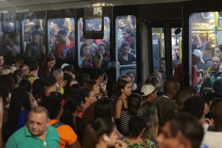 Na imagem, diversas pessoas na fila de um ponto de ônibus