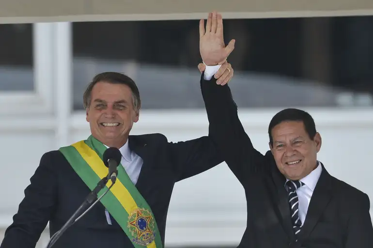 Dois homens brancos sorrindo de terno levantando as mãos