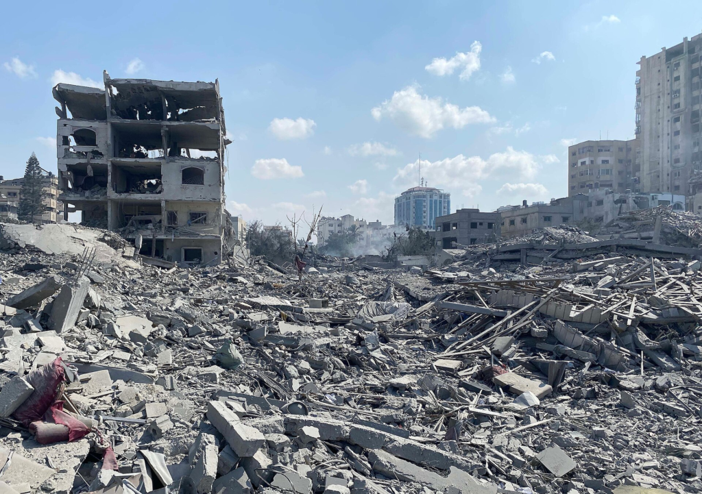 Fotografia em plano geral e iluminação natural de uma área urbana devastada por escombros de construções destruídas. O primeiro plano é dominado por uma grande quantidade de entulho cinzento, composto por pedaços de concreto, vigas de ferro e fragmentos de madeira espalhados. À esquerda, destaca-se a estrutura parcial de um prédio de quatro andares severamente danificado, com paredes destruídas e interior exposto. Ao fundo, sob um céu claro com poucas nuvens, veem-se outros edifícios distantes e uma torre moderna em tons azuis que permanece de pé.