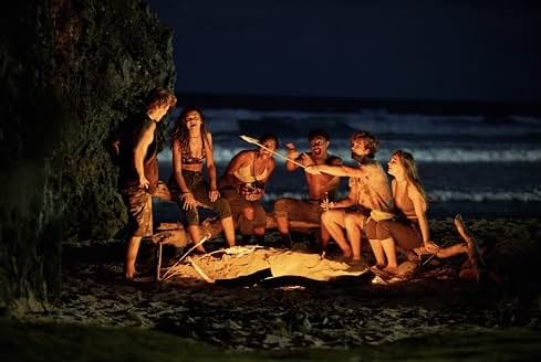 Fotografia noturna em plano geral de um grupo de seis jovens reunidos ao redor de uma fogueira acesa na areia de uma praia, com ondas do mar quebrando ao fundo sob a escuridão do céu. No canto esquerdo, um paredão rochoso escuro delimita o espaço onde o grupo está. Um rapaz de bermuda permanece em pé à esquerda, enquanto os outros cinco amigos estão assentados em troncos de madeira e na areia ao redor do fogo, rindo e interagindo descontraídos. A luz alaranjada das chamas ilumina o grupo no centro, destacando um rapaz sem camisa apontando uma vara de madeira em direção ao fogo enquanto os amigos observam e se divertem.