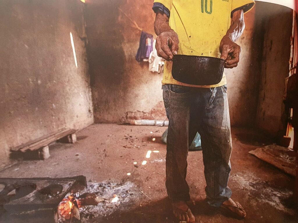 Fotografia em plano médio de uma pessoa em pé dentro de um cômodo humilde de chão de terra batida e paredes de alvenaria inacabadas. A pessoa é enquadrada do pescoço para baixo e veste uma camiseta amarela da seleção brasileira de futebol, com o número dez verde no peito, e calça jeans surrada. Com as duas mãos calejadas, ela segura à frente do corpo uma pequena panela preta de ferro encharcada de fuligem. No canto inferior esquerdo, sobre o chão de terra, há um fogão a lenha improvisado com chamas alaranjadas acesas. Ao fundo, o ambiente rústico possui pouca mobília, destacando-se um pequeno banco de madeira encostado na parede e roupas penduradas.