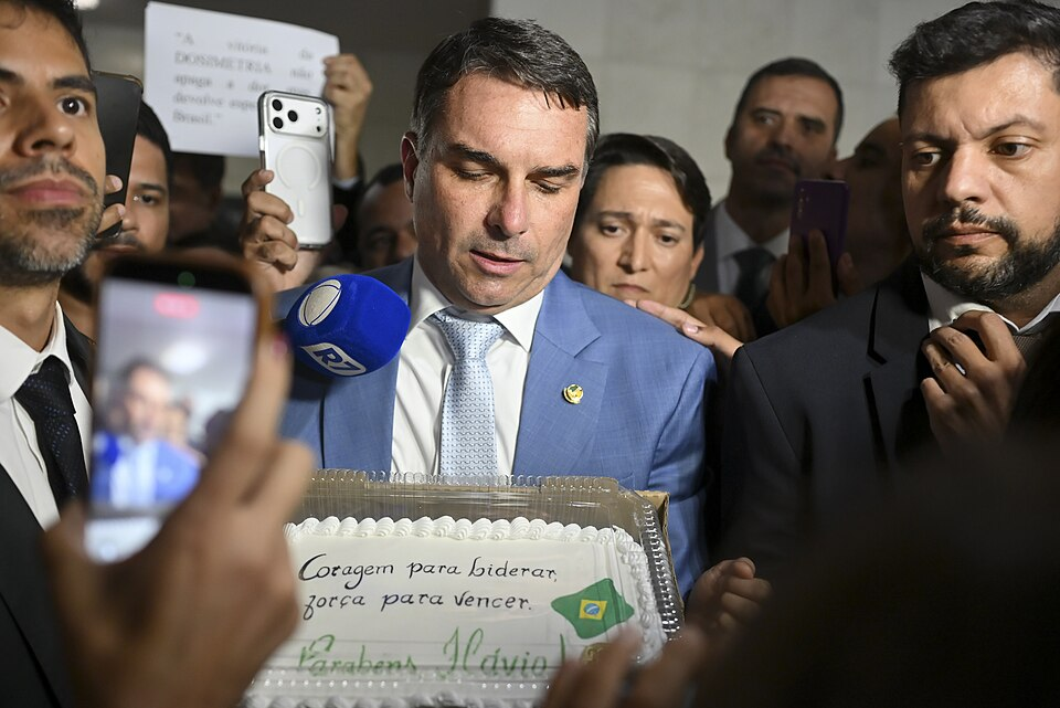 Homem branco de terno azul segurando um bolo na frente de jornalistas