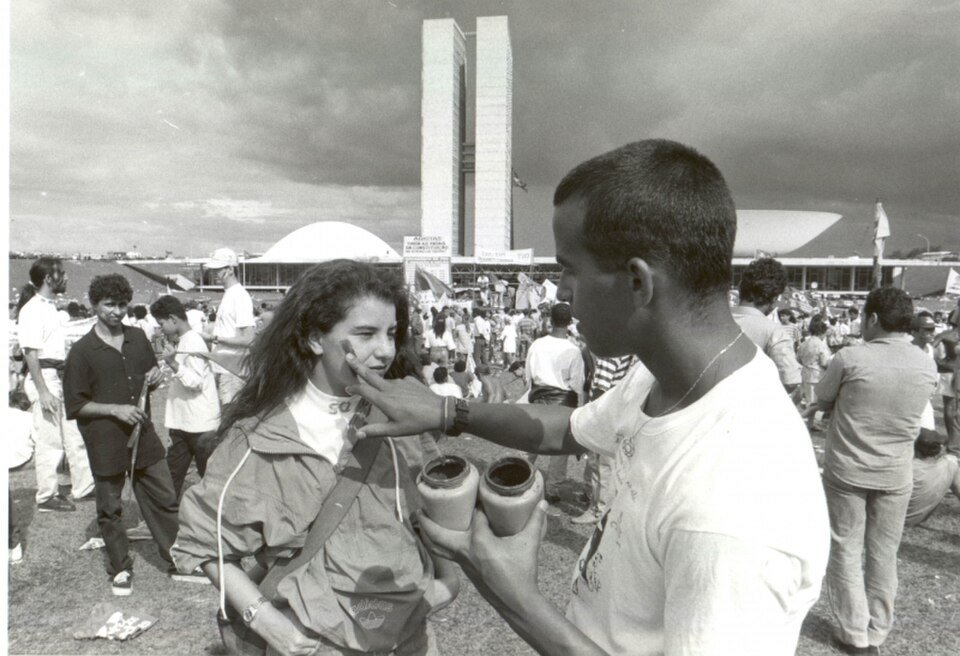 Homem pardo de camisa branca pintando o rosto de uma mulher branca e de cabelos longos