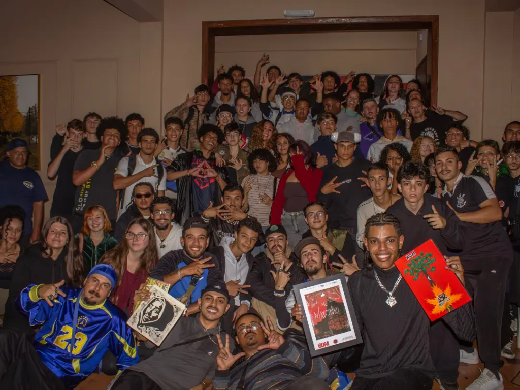 Fotografia em plano geral e ângulo frontal mostrando um grande grupo de jovens reunidos em um saguão interno. Eles estão agrupados em vários níveis — alguns sentados no chão à frente e outros em pé ao fundo —, sorrindo e fazendo gestos com as mãos para a câmera. Vários deles vestem casacos esportivos e seguram quadros e encartes artísticos nas mãos.