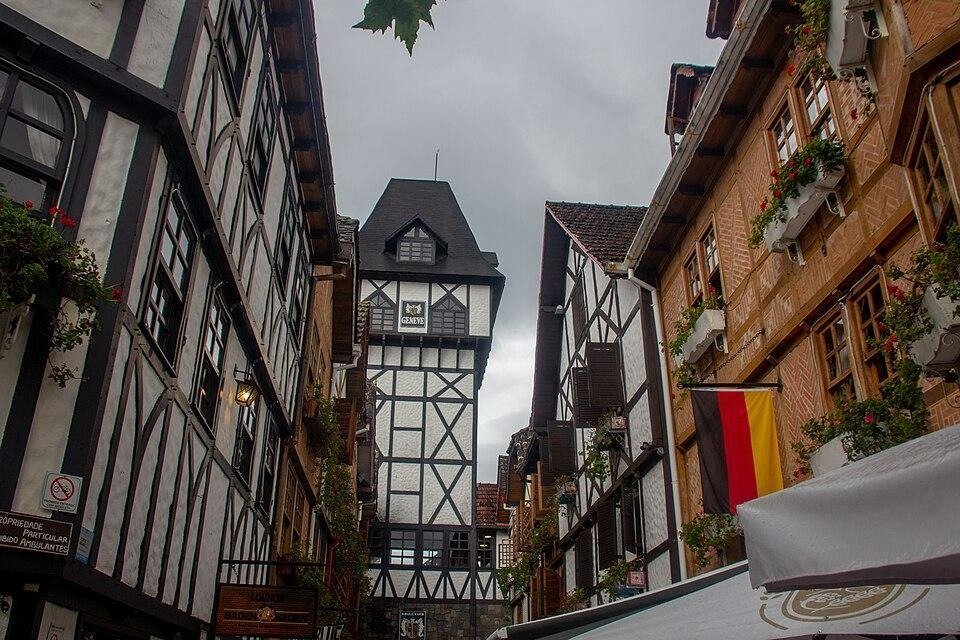 Fotografia de rua em ângulo baixo mostrando uma ruela estreita com arquitetura estilo enxaimel. Os prédios altos das laterais possuem fachadas brancas com vigas de madeira escura cruzadas, janelas floreiras e lâmpadas de estilo antigo. Ao fundo, destaca-se uma torre com o letreiro "GENEVE", enquanto a bandeira da Alemanha aparece pendurada na fachada do prédio à direita, sob um céu nublado.
